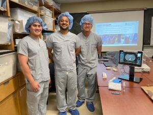 Three surgical staff members stand in a lab wearing scrubs, hair nets, and shoe covers during a hands-on demo of NZTech’s touchless OR technology. A medical display beside them shows an image from the TIPSO AirPad™ system.
