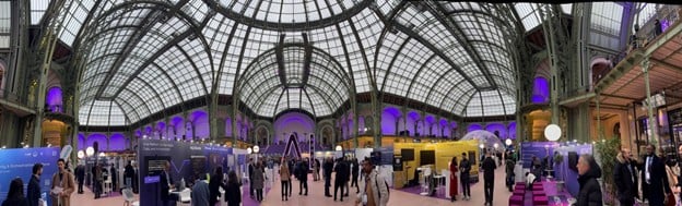 Panoramic view of the Adopt AI Summit inside the Grand Palais in Paris, showing exhibitor booths, attendees, and the historic glass-domed architecture illuminated with purple lighting.