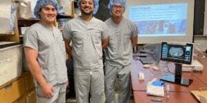 Three surgical staff members stand in a lab wearing scrubs, hair nets, and shoe covers during a hands-on demo of NZTech’s touchless OR technology. A medical display beside them shows an image from the TIPSO AirPad™ system.